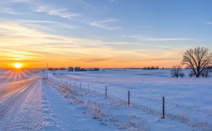 Snow-covered Kansas rural road and prairie at sunrise with farm buildings in the distance.